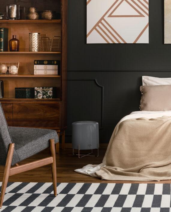 Lifestyle image of Vornado CYLO51 Air Purifier with True HEPA Filtration on wood floor between a bed and a tall bookshelf.