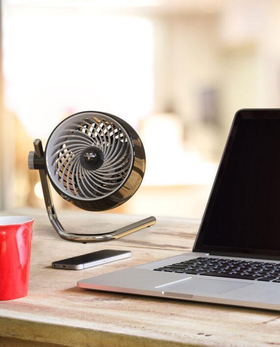 Lifestyle of a black Pivot3 compact air circulator on a desk by a phone and an opened laptop