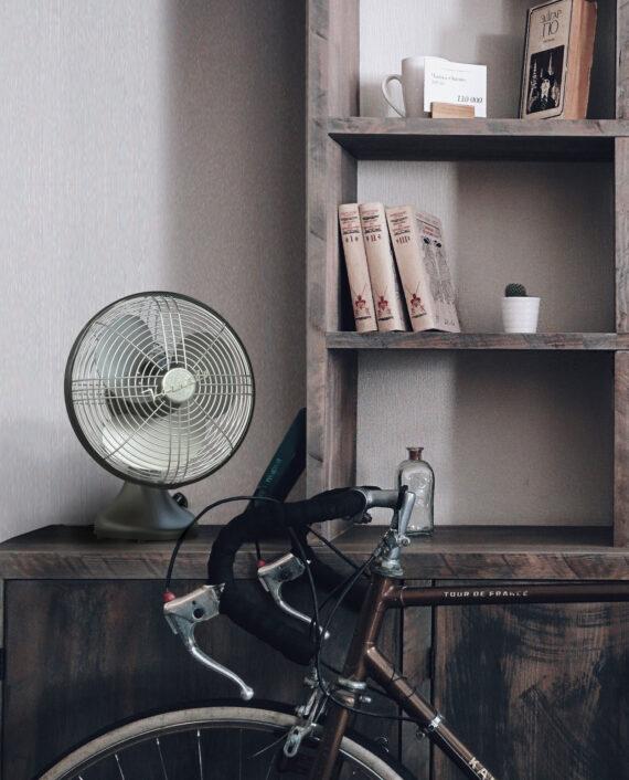 Lifestyle of a Matte Black Silver Swan S Vintage Oscillating fan on a wooden cabinets with a bicycle leaning next to it